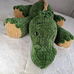 Könnte beinhalten: Ein grünes Dinosaurier-Plüschtier mit beigen Akzenten an Füßen und Rücken. Das handgefertigte Stofftier hat schwarze Augen und liegt auf einer weißen Oberfläche. Das weiche, strukturierte Material verleiht dem Spielzeug ein kuscheliges Aussehen.