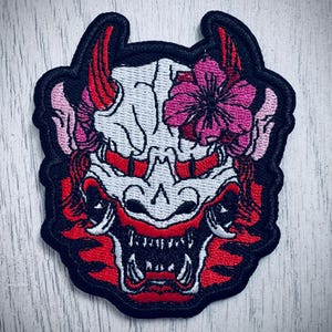 May include: Embroidered patch featuring a stylized oni mask design. The mask is primarily white with red accents, including horns, mouth, and details. Pink cherry blossoms adorn the top. The patch has a black border.