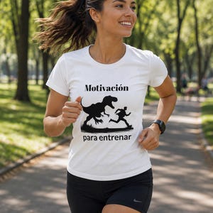 Puede incluir: Una camiseta blanca de manga corta con un estampado gráfico negro de un dinosaurio persiguiendo a una persona. El texto "Motivación" aparece sobre el gráfico y el texto "para entrenar" aparece debajo del gráfico en una fuente negra.