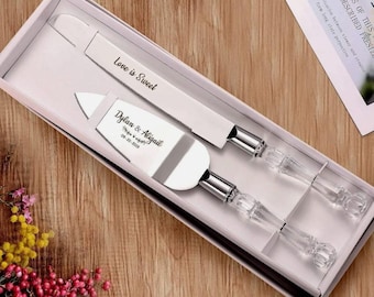 May include: A wedding cake knife and server set in a white box. The knife has the text "Love is Sweet" and the server has the names "Dylan & Abigail" and the date "09-20-2019". The handles are clear with a crystal-like design.