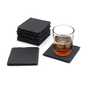 May include: A set of square black slate coasters with rough, natural edges. One coaster is placed separately, one is positioned under a clear glass tumbler filled with an amber-coloured liquid and ice cubes, and a stack of six coasters sits nearby.