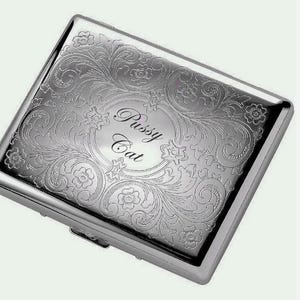 May include: A rectangular silver-toned metal cigarette case featuring an intricate engraved floral and scrollwork pattern across the surface. The center of the case displays an oval frame containing the engraved text Pussy Cat in a script font.