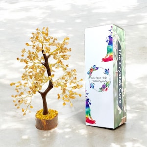 Könnte beinhalten: Eine dekorative Baumskulptur mit braunem Stamm und Ästen, die mit gelben Kristallen verziert sind. Der Baum steht auf einem Holzsockel. Daneben befindet sich eine Schachtel mit dem Text "The Crystal Cure" und einer farbenfrohen Illustration.