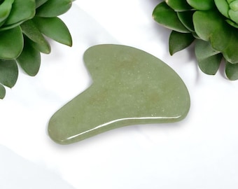 May include: A light green jade gua sha tool with a smooth, rounded surface. The tool is shaped like a crescent moon and is used for facial massage.
