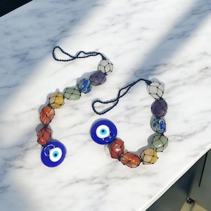 May include: Two decorative hanging ornaments. Each features a string of colorful stones in a net, with a blue glass eye charm. The stones are in shades of red, orange, yellow, green, blue, purple, and white. The background is a white marble surface.