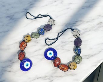 Puede incluir: Dos adornos colgantes decorativos. Cada uno presenta una cadena de piedras de colores en una red, con un amuleto de ojo de vidrio azul. Las piedras son en tonos de rojo, naranja, amarillo, verde, azul, morado y blanco. El fondo es una superficie de mármol blanco.