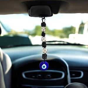 Può includere: Un ciondolo per auto appeso allo specchietto retrovisore. Il ciondolo presenta una fila di pietre bianche e nere, sormontata da una perla a forma di occhio blu e bianco. Il ciondolo è sospeso davanti al cruscotto di un'auto.