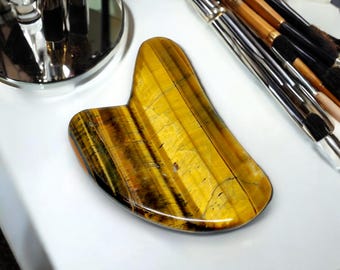 May include: A tiger's eye gua sha tool, a smooth, heart-shaped stone with a golden yellow, brown, and black pattern. The tool is used for facial massage and lymphatic drainage.