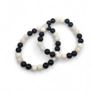 Puede incluir: Dos pulseras hechas de cuentas negras y blancas. Las pulseras están hechas de cuentas redondas y están apiladas una encima de la otra.