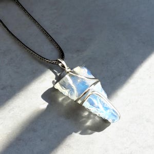 May include: A silver wire wrapped pendant featuring a clear, iridescent, blue and white crystal. The pendant is attached to a black cord.