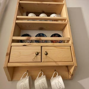 Peut inclure: Une étagère murale en bois avec deux tiroirs et des crochets pour accrocher des tasses. L'étagère a trois tablettes pour exposer des objets. Il y a trois tasses blanches accrochées aux crochets et plusieurs bols décoratifs sur l'étagère supérieure.
