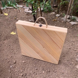 May include: A wooden folding table with a diagonal wood grain pattern. The table is closed and has a rope handle.