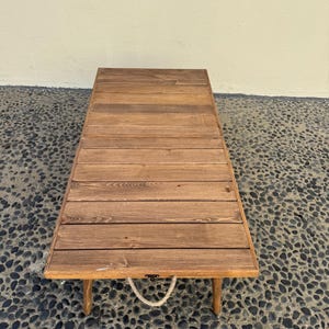 May include: A wooden folding table with a rope handle. The table is made of natural wood and has a rustic look. It is sitting on a bed of small black stones.