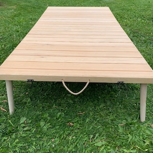 May include: A light-colored wooden picnic table with a slatted top and four short, rounded legs. The table has a rope handle and metal hinges. The table is set on a bed of green grass.