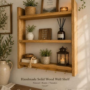 Puede incluir: Un estante de pared de madera maciza hecho a mano con tres niveles y cinco ganchos de metal. El estante mide 60 cm de ancho, 40 cm de alto y 9 cm de profundidad. Está decorado con pequeñas plantas en macetas, un cuadro, una vela y un farol.