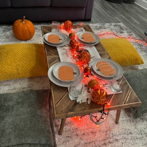 May include: A rustic wooden table with a white tablecloth and string lights. There are four place settings with orange pumpkin-shaped plates and glasses. The table is surrounded by pillows and a pumpkin.
