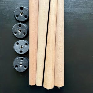 May include: A set of four unfinished wooden furniture legs with a tapered design, alongside four black plastic mounting plates. The legs are light brown and appear ready for staining or painting. Hardware is included.