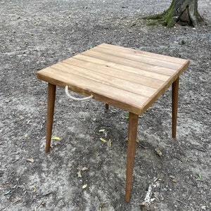 Pode incluir: Uma mesa quadrada de madeira com uma alça de corda na lateral. A mesa é feita de madeira marrom clara e tem um acabamento natural. A mesa está situada em um caminho de terra em uma floresta.