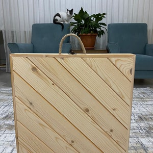 May include: A wooden box with a diagonal wood grain pattern and a rope handle. The box is closed and sitting on a patterned rug.