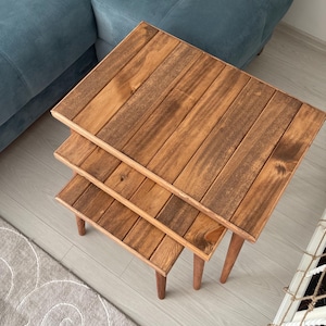 Puede incluir: Juego de tres mesas nido de madera en color marrón cálido. Cada mesa, de forma cuadrada, está hecha de tablones de madera y tiene patas redondeadas. Las mesas están dispuestas en una configuración apilada, con la más pequeña en la base.