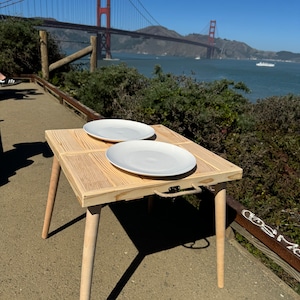 May include: A wooden folding table with two white plates on top. The table has a rope handle and is set up outdoors with a view of the Golden Gate Bridge in the background.