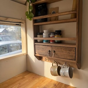 May include: A rustic wooden shelf unit with multiple shelves, a drawer, and hooks. The shelves hold jars, containers, and coffee bags. Two ceramic mugs hang from the hooks. The unit is mounted on a light-colored wall, with a window visible on the left.