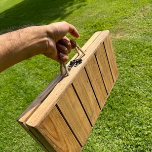 Puede incluir: Una caja de madera con un asa de cuerda y un cierre de metal. La caja está hecha de madera marrón claro y tiene un aspecto rústico.