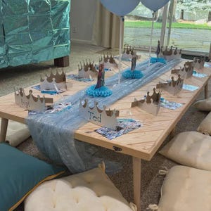 Peut inclure: Une table en bois décorée pour une fête, avec un chemin de table bleu clair et des couronnes en carton avec des noms comme "Gigi" et "Ted". Des ballons bleus et des figurines décoratives sont également présents. Des coussins de sol entourent la table.