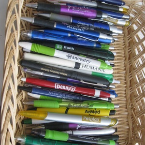 May include: A wicker basket filled with a variety of pens. The pens are in various colors, including blue, green, red, and black, and many have company names and logos printed on them. The basket is made of woven light brown wicker.