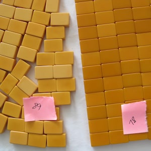 May include: A collection of rectangular mustard yellow tiles arranged in two groups. One group is organized in a neat grid, while the other is scattered. Two small pink square notes are placed on the tiles, with the handwritten numbers 89 and 78.