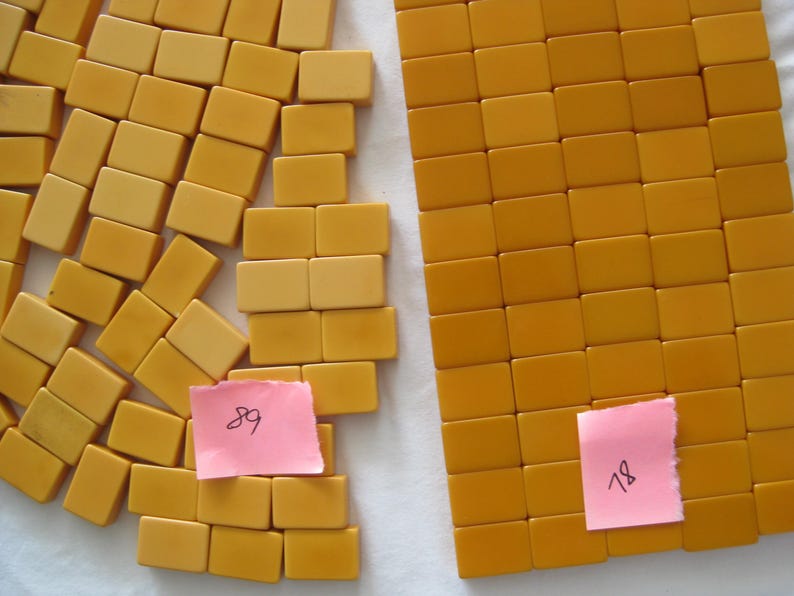 May include: A collection of rectangular mustard yellow tiles arranged in two groups. One group is organized in a neat grid, while the other is scattered. Two small pink square notes are placed on the tiles, with the handwritten numbers 89 and 78.