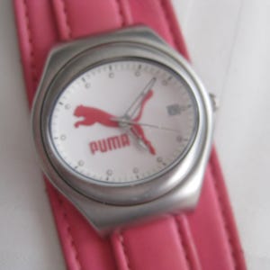 K&ouml;nnte beinhalten: Eine silberne Armbanduhr mit wei&szlig;em Zifferblatt und einem rosafarbenen Lederarmband. Das Zifferblatt der Uhr zeigt ein rotes Puma-Logo und darunter den Schriftzug "PUMA".