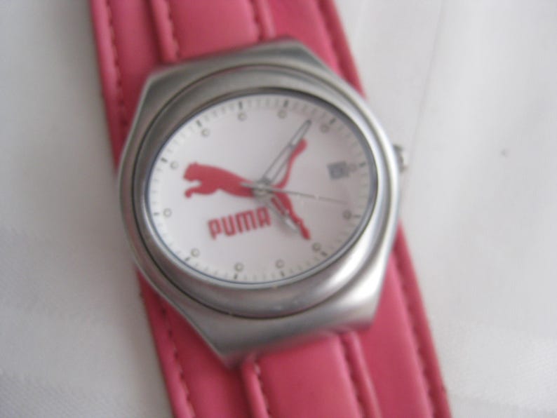 K&ouml;nnte beinhalten: Eine silberne Armbanduhr mit wei&szlig;em Zifferblatt und einem rosafarbenen Lederarmband. Das Zifferblatt der Uhr zeigt ein rotes Puma-Logo und darunter den Schriftzug "PUMA".