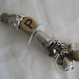 May include: A silver charm bracelet with a wooden letter P charm, a thimble, a silver chain, and various other charms, including a clear crystal, a silver heart charm, and a brown bead.