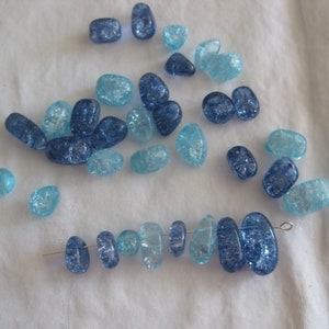 May include: A collection of blue and light blue glass beads with a crackle effect. Some beads are scattered, while others are strung on a wire, showcasing their varying shapes and sizes. The beads are on a white fabric background.