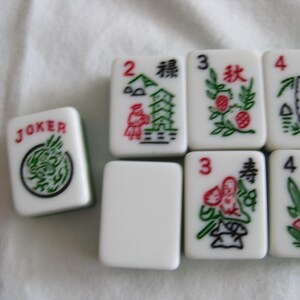 K&ouml;nnte beinhalten: Eine Sammlung wei&szlig;er Mahjong-Steine mit roten, gr&uuml;nen und schwarzen Symbolen und Zeichen. Ein Stein tr&auml;gt die Aufschrift "JOKER" mit einem gr&uuml;nen Drachendesign. Andere Steine zeigen Zahlen und Illustrationen von Pflanzen und Geb&auml;uden.