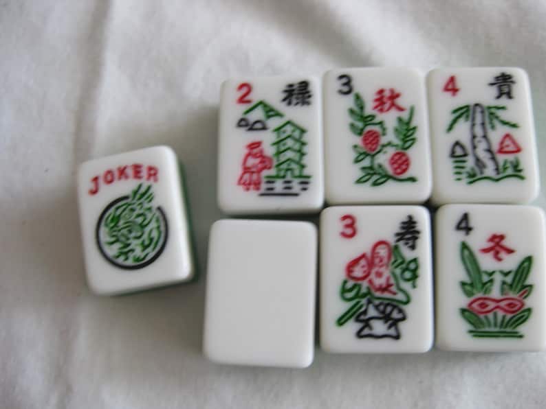 K&ouml;nnte beinhalten: Eine Sammlung wei&szlig;er Mahjong-Steine mit roten, gr&uuml;nen und schwarzen Symbolen und Zeichen. Ein Stein tr&auml;gt die Aufschrift "JOKER" mit einem gr&uuml;nen Drachendesign. Andere Steine zeigen Zahlen und Illustrationen von Pflanzen und Geb&auml;uden.