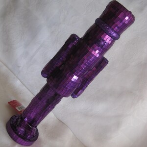 May include: A nutcracker figurine covered in small, reflective purple square mirror tiles. The figure features a cylindrical hat, a uniform-style torso with arms at the sides, and a circular base. A small red tag is attached to the lower section.