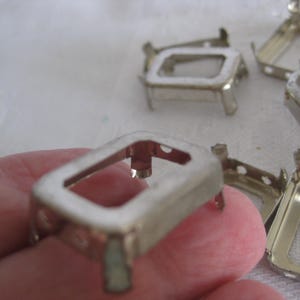May include: A collection of silver-toned metal rectangular settings featuring four small prongs at each corner. These open-frame components have a hollow centre and are designed for mounting small items, with several pieces arranged in a group.