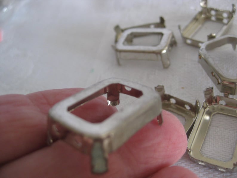 May include: A collection of silver-toned metal rectangular settings featuring four small prongs at each corner. These open-frame components have a hollow centre and are designed for mounting small items, with several pieces arranged in a group.