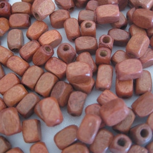 May include: A collection of small, reddish-brown wooden beads. The beads are rectangular and cylindrical, with a hole running through the center of each. They are scattered across a light-colored surface.