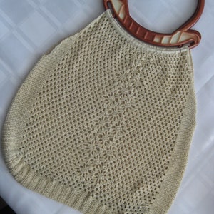 May include: A vintage, cream-colored crocheted handbag with a decorative floral pattern. The bag features a sturdy, brown and cream-colored handle. The open-weave design is visible throughout the bag, showcasing the intricate crochet work.