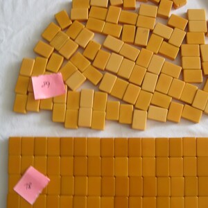 May include: A collection of rectangular, mustard-yellow mahjong tiles arranged in a large, loose cluster and a neat, organized rectangular grid. Two small, pink adhesive notes with handwritten numbers are attached to the tile groups.