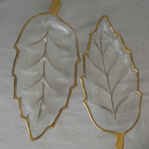 May include: Two clear glass leaf-shaped serving dishes with gold trim. The dishes are empty and are sitting on a white surface.