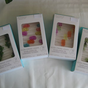 May include: Four boxes of Ashland brand string lights with 18 lights each. The lights are shaped like cacti, popsicles, and palm trees. Each box has a white background with blue and green accents. The boxes are labeled "ICON STRING LIGHTS" and "GUIRLANDE DE LUMIÈRES ICONES".