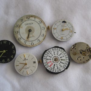 May include: A collection of vintage watch faces and movements, including a black face with gold hands, a white face with the brand "Orien", and a face with a calendar dial. The watch faces are in various states of wear.