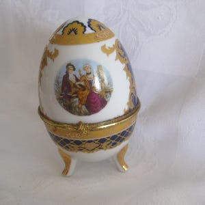 May include: A decorative egg-shaped trinket box with a hinged lid. The white porcelain egg features gold accents, blue diamond patterns, and a painted scene of two figures. The egg rests on three gold-tipped feet.