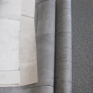 May include: Close-up of several rolls of textured wallpaper in shades of gray and beige. The wallpaper has a brick-like pattern and a subtle, mottled appearance. The rolls are partially unrolled, revealing the different textures and colors.