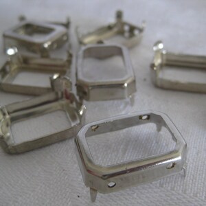 May include: A collection of silver-toned metal rectangular settings with prongs. Each piece features a hollow centre and small holes on the sides, designed for mounting stones or cabochons. The metallic finish is reflective and smooth.