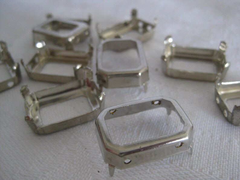 May include: A collection of silver-toned metal rectangular settings with prongs. Each piece features a hollow centre and small holes on the sides, designed for mounting stones or cabochons. The metallic finish is reflective and smooth.